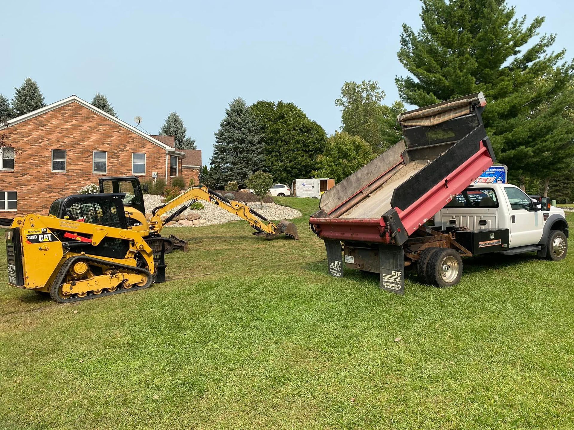 Two work trucks in a grassy yard, one dumping debris from a raised bed near a brick house.