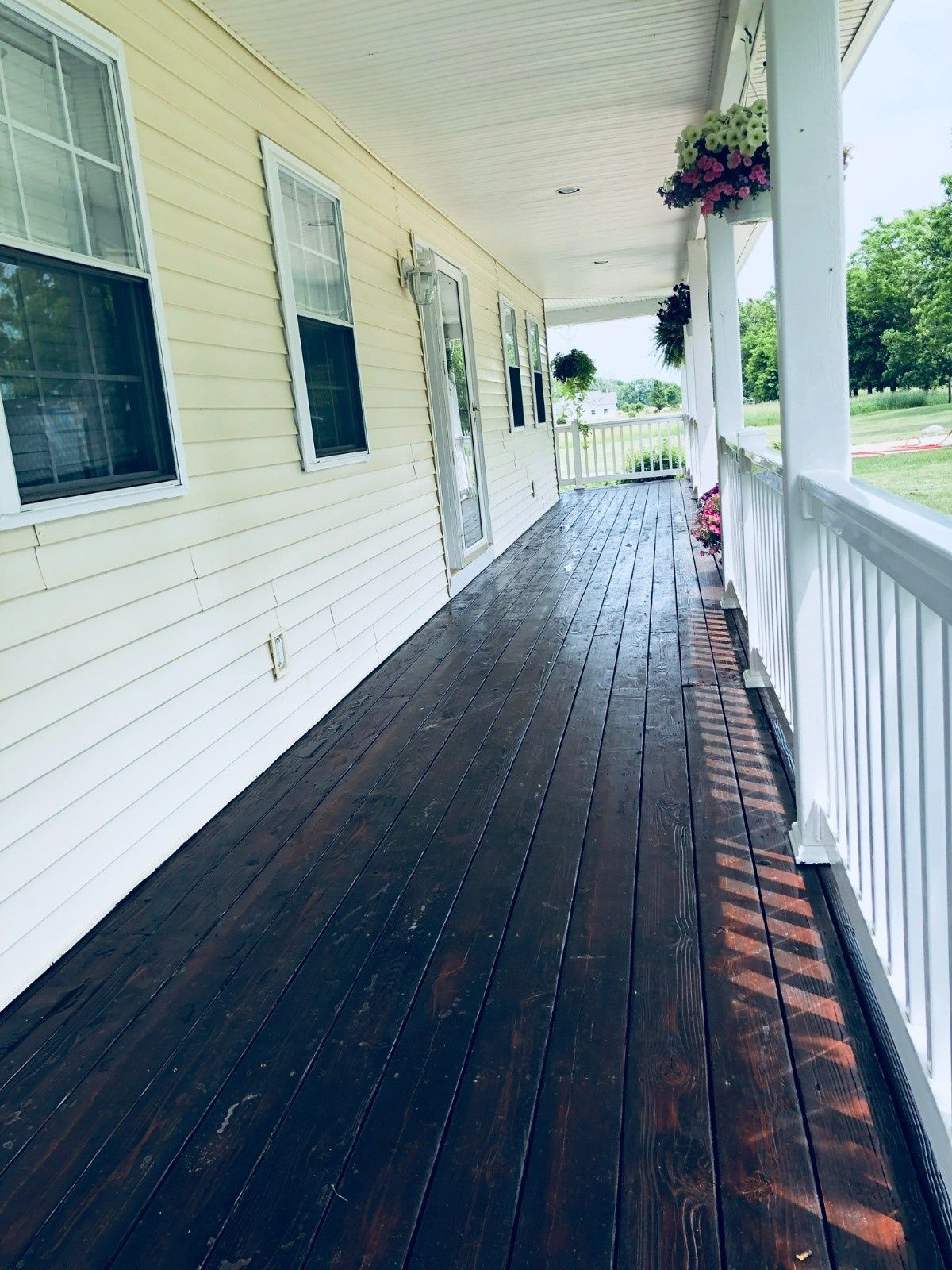 Covered porch with dark wooden floor, white siding, windows, and hanging flowers overlooking a yard
