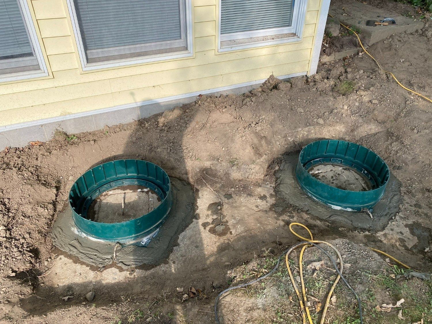 Two green pipe rings set in dirt beside a house foundation and windows.