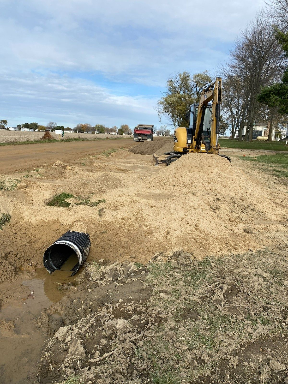 Excavator digging a trench beside a dirt road, with a pipe outlet in the foreground and a truck in the distance.