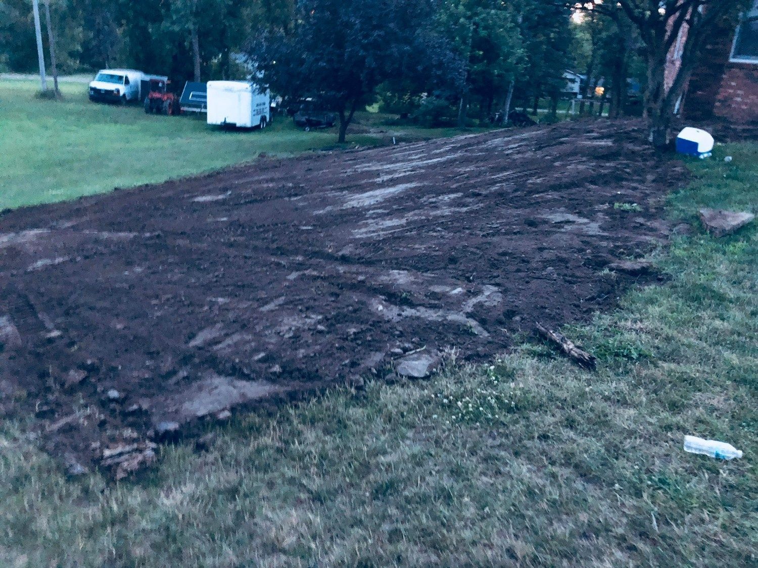 Muddy dirt clearing in a grassy yard with trees and a few parked vehicles in the background