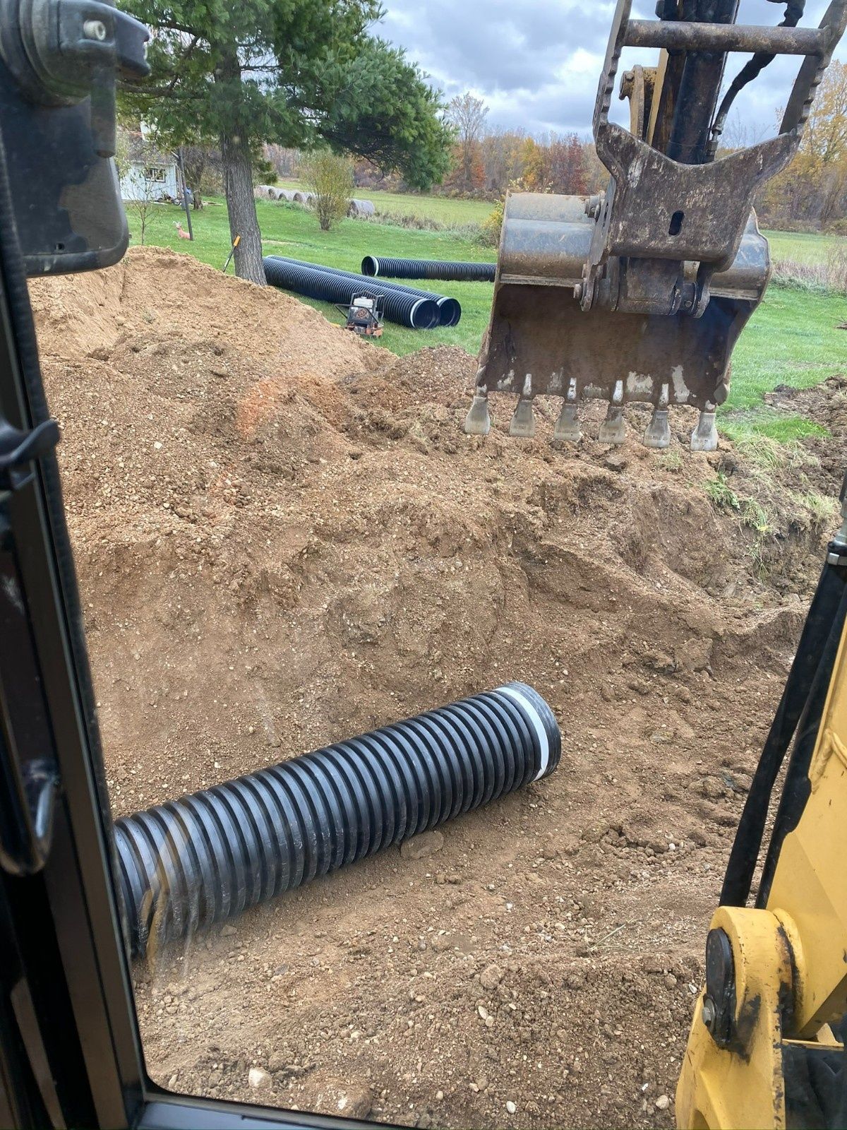 Excavator bucket above a trench with black corrugated pipes and dirt at a construction site