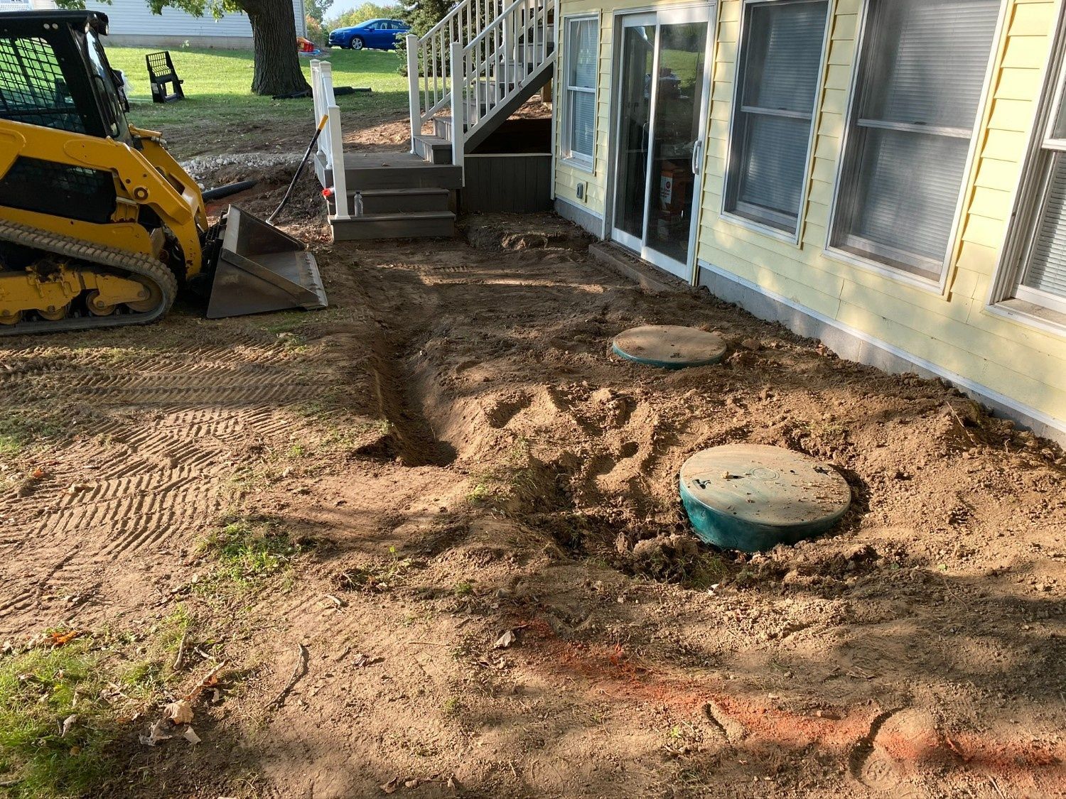 Backyard construction site with trench, excavator, and dirt beside a house.