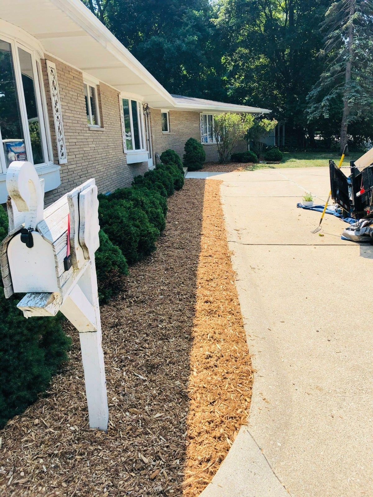 Brick house driveway with mulch border, white mailbox, and lawn equipment near the garage.