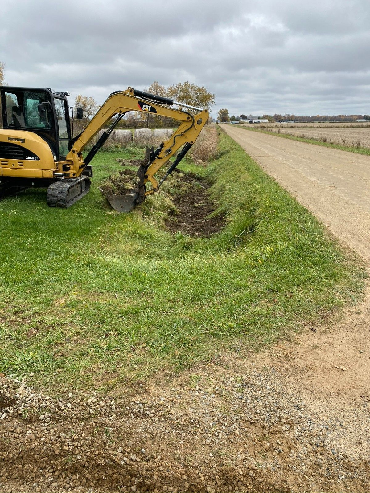 Yellow excavator digging a ditch beside a grassy field and gravel road