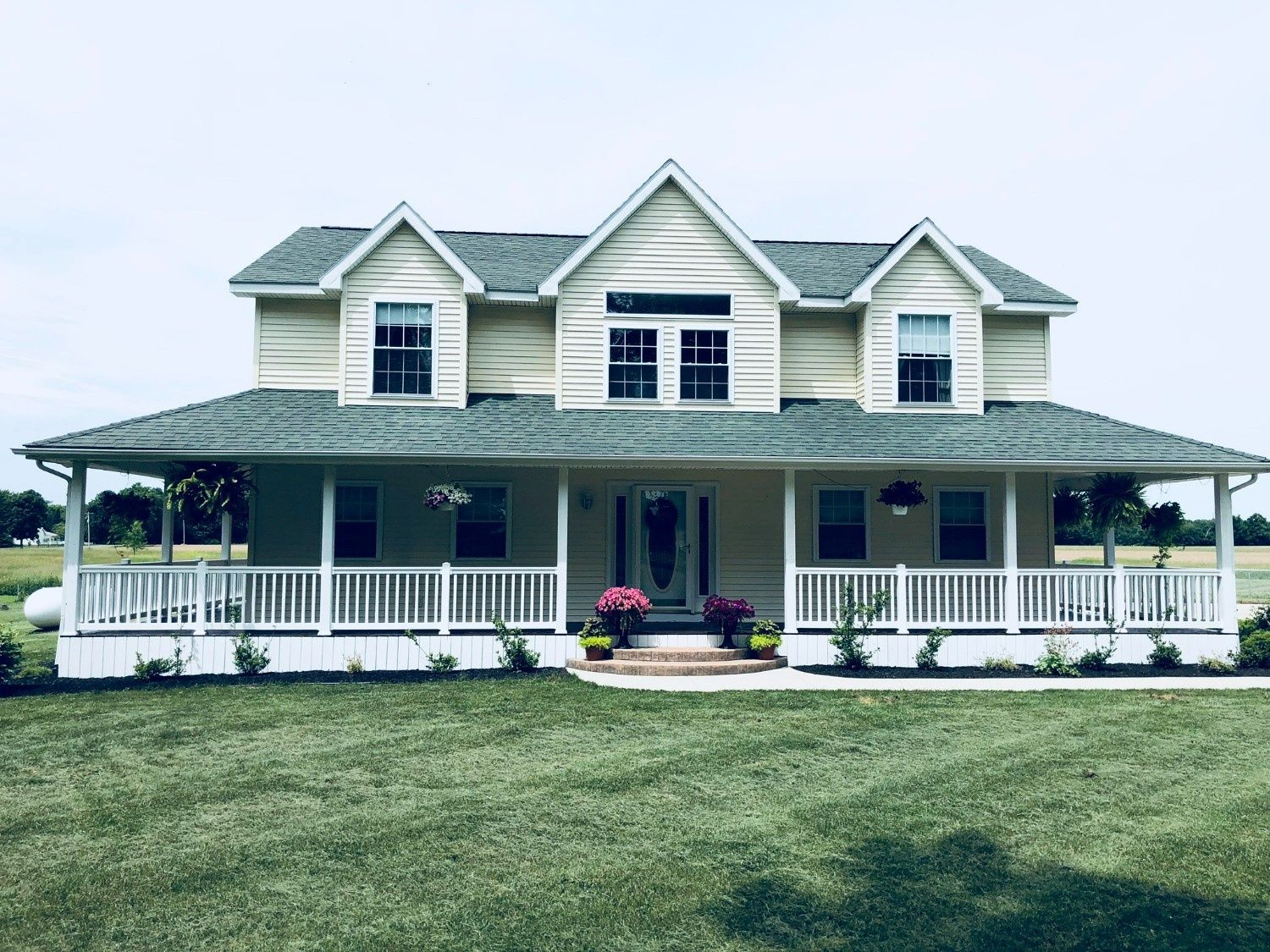 White two-story farmhouse with wraparound porch and lawn under a cloudy sky