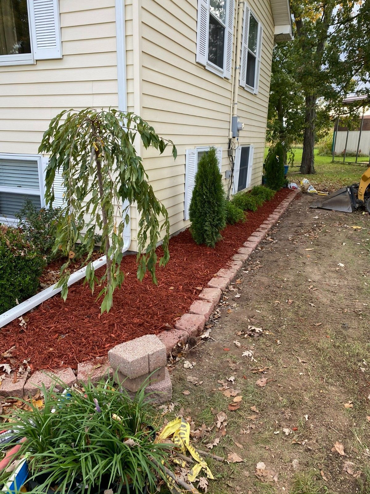 Side yard with beige house, red mulch border, shrubs, and a small tree along a brick edging.