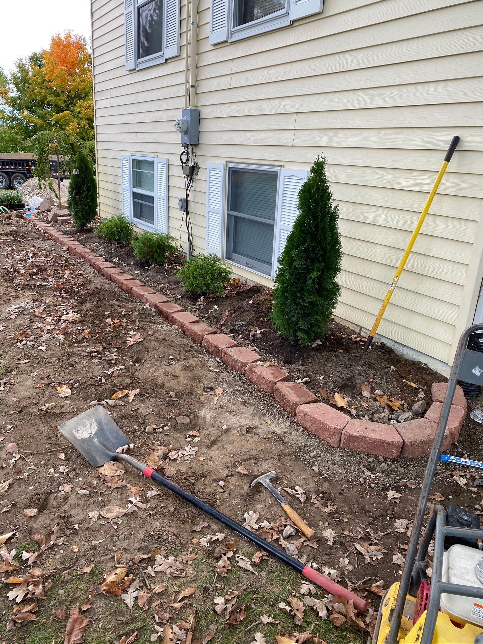 Side of a beige house with planted shrubs, a shovel on dirt, and fall leaves along the foundation