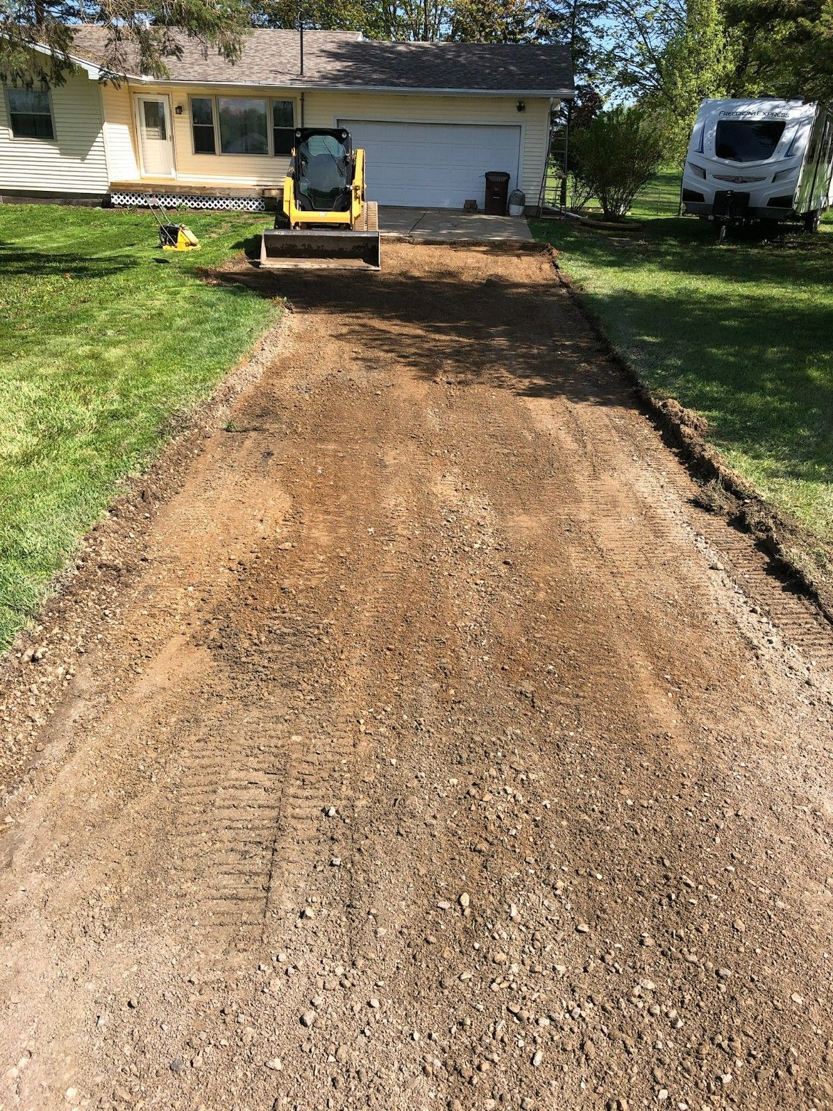Small excavator leveling a long dirt driveway in front of a house, with a truck parked nearby.