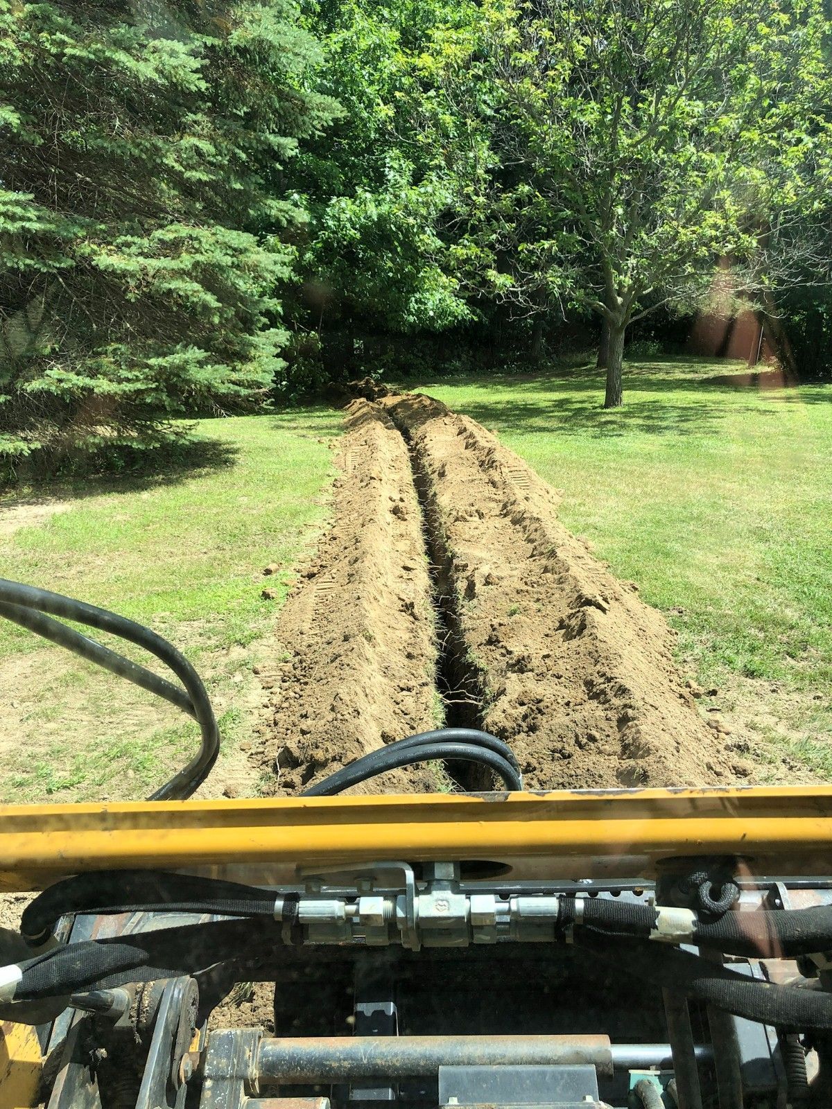 View from a tractor digging a long trench through a grassy yard with trees nearby.