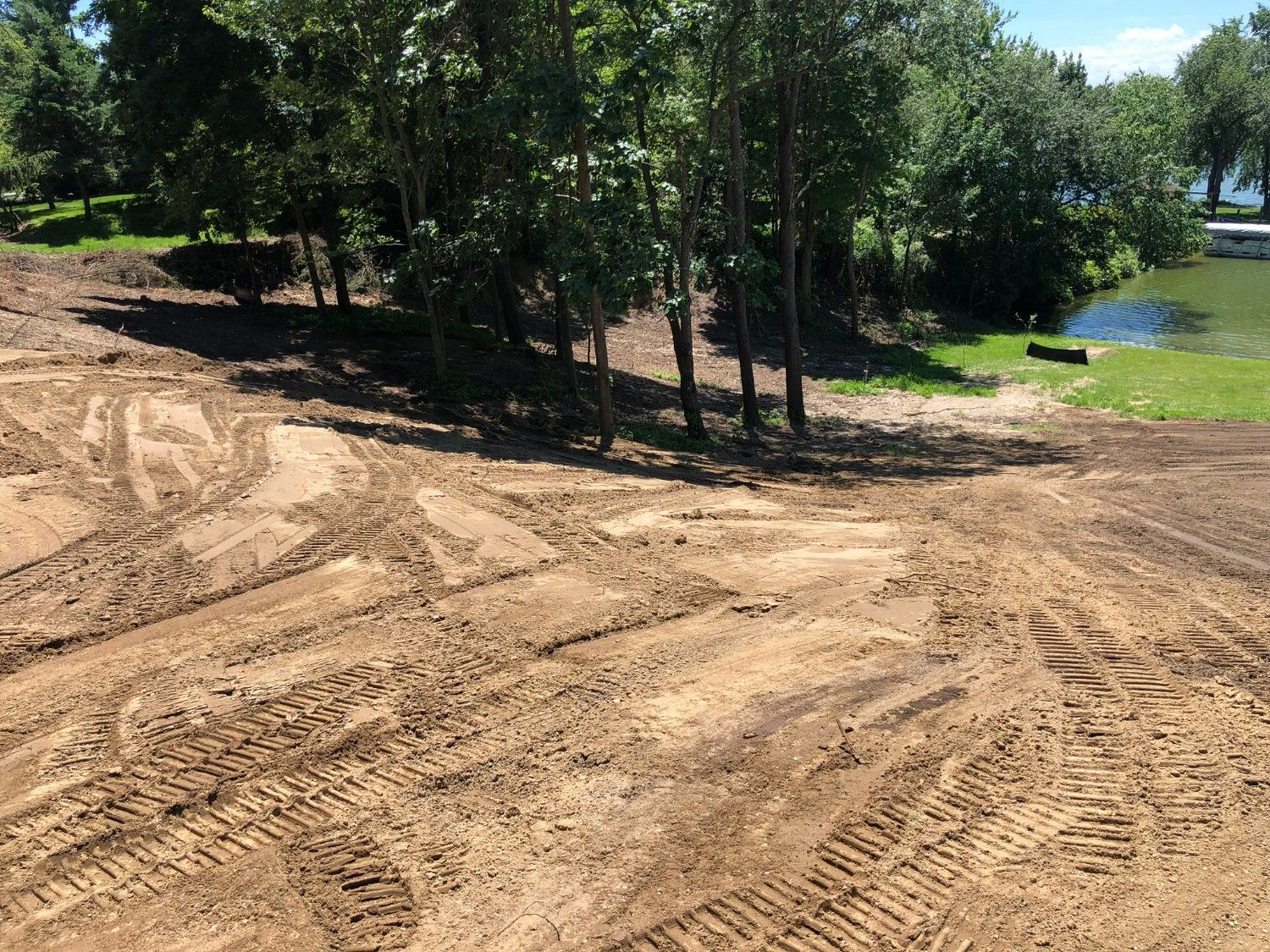 Freshly graded dirt path beside trees and a grassy pond under bright sunlight