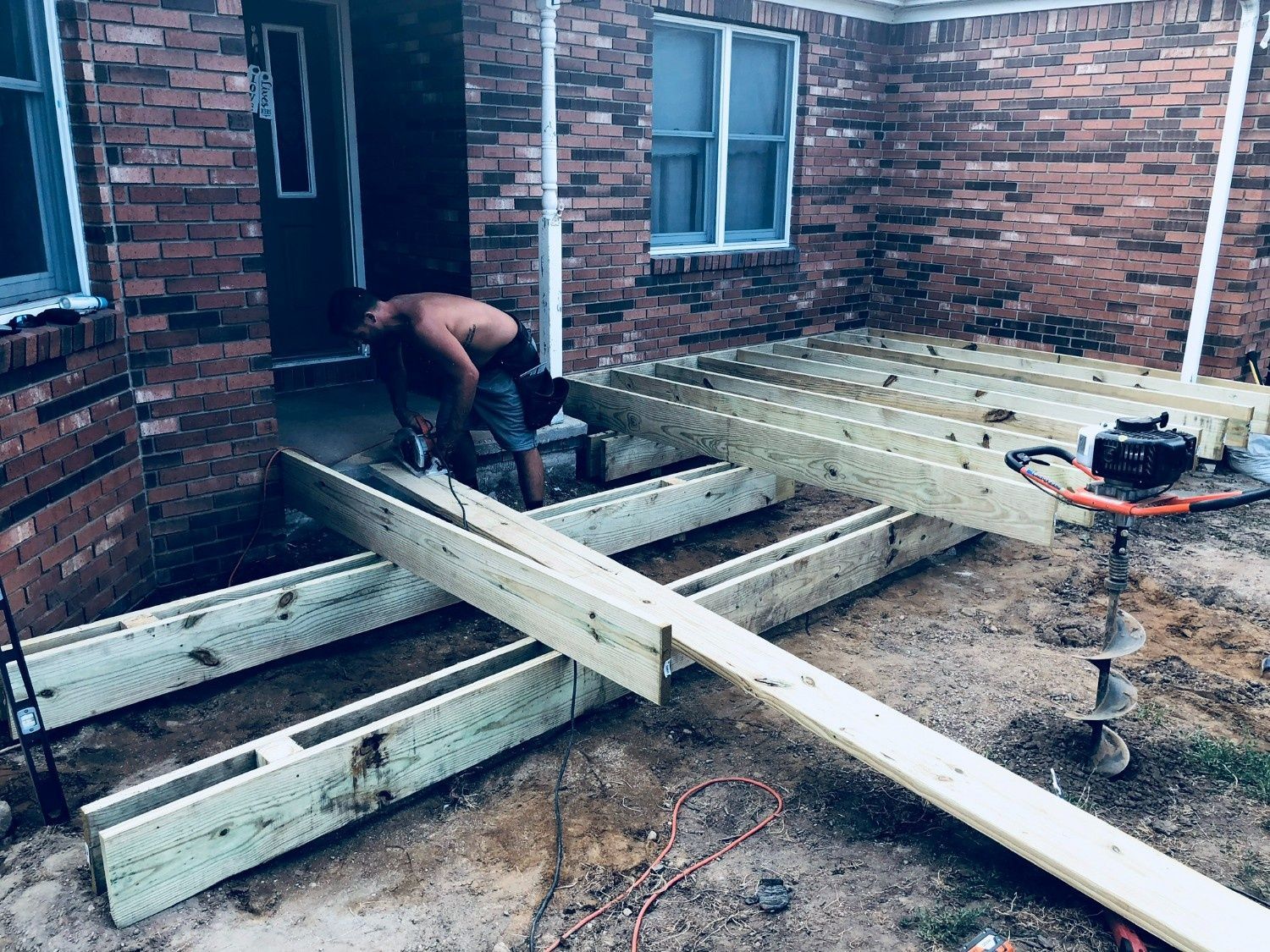 Worker building wooden steps beside a brick house, with lumber and tools on the ground