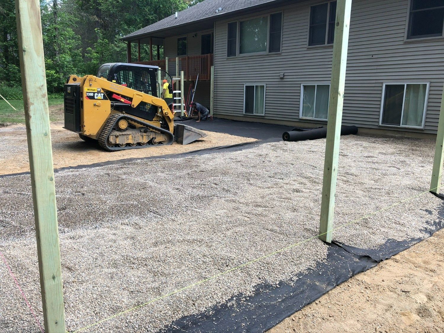 Yellow skid steer leveling gravel beside a gray house under construction, with green support posts in front.