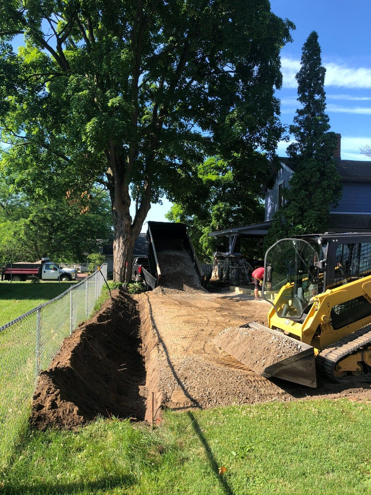 Construction equipment leveling a dirt path beside a fenced lawn with trees and a house.
