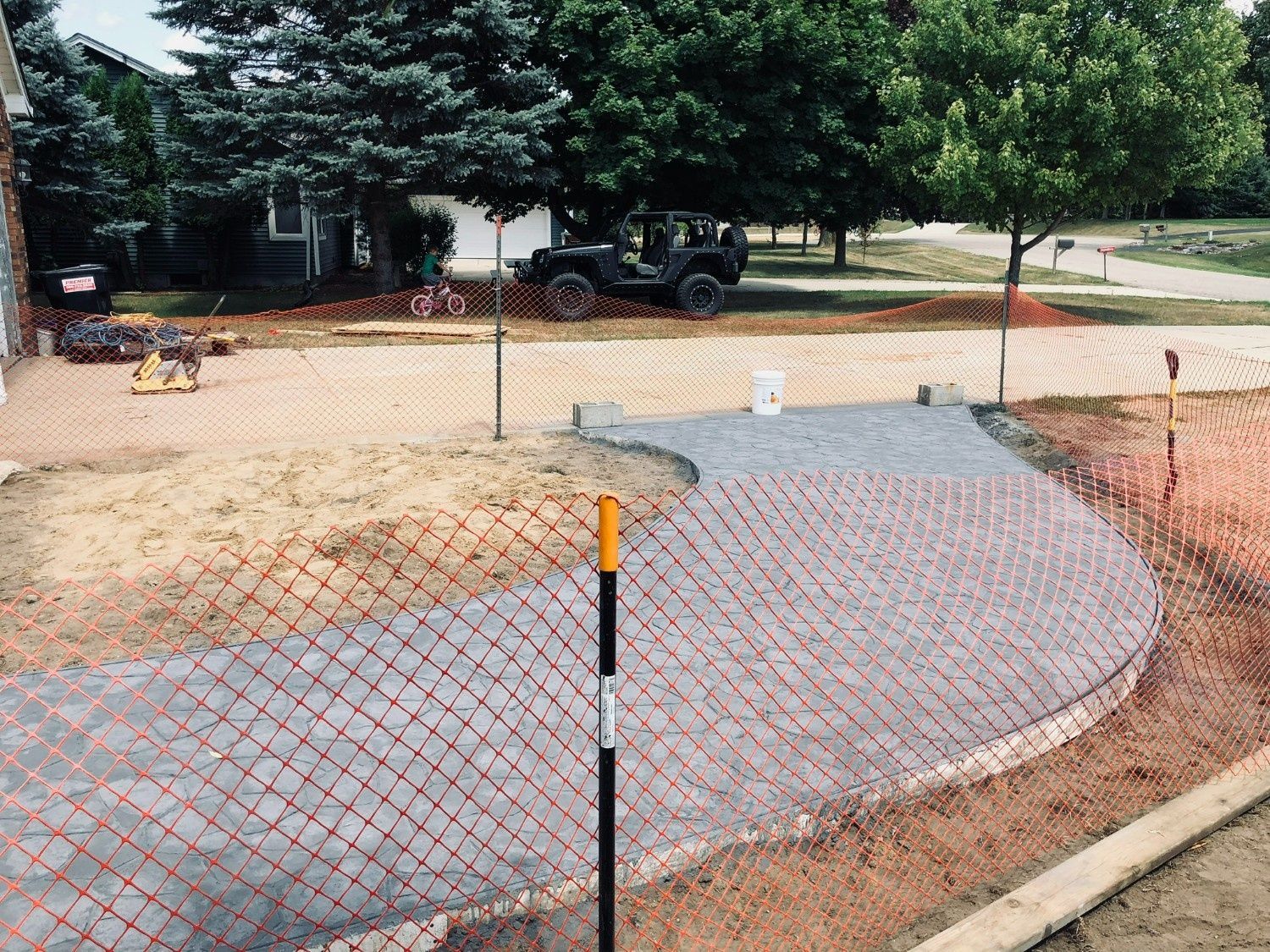 Curved brick walkway under construction with orange safety netting and trees in a park-like area
