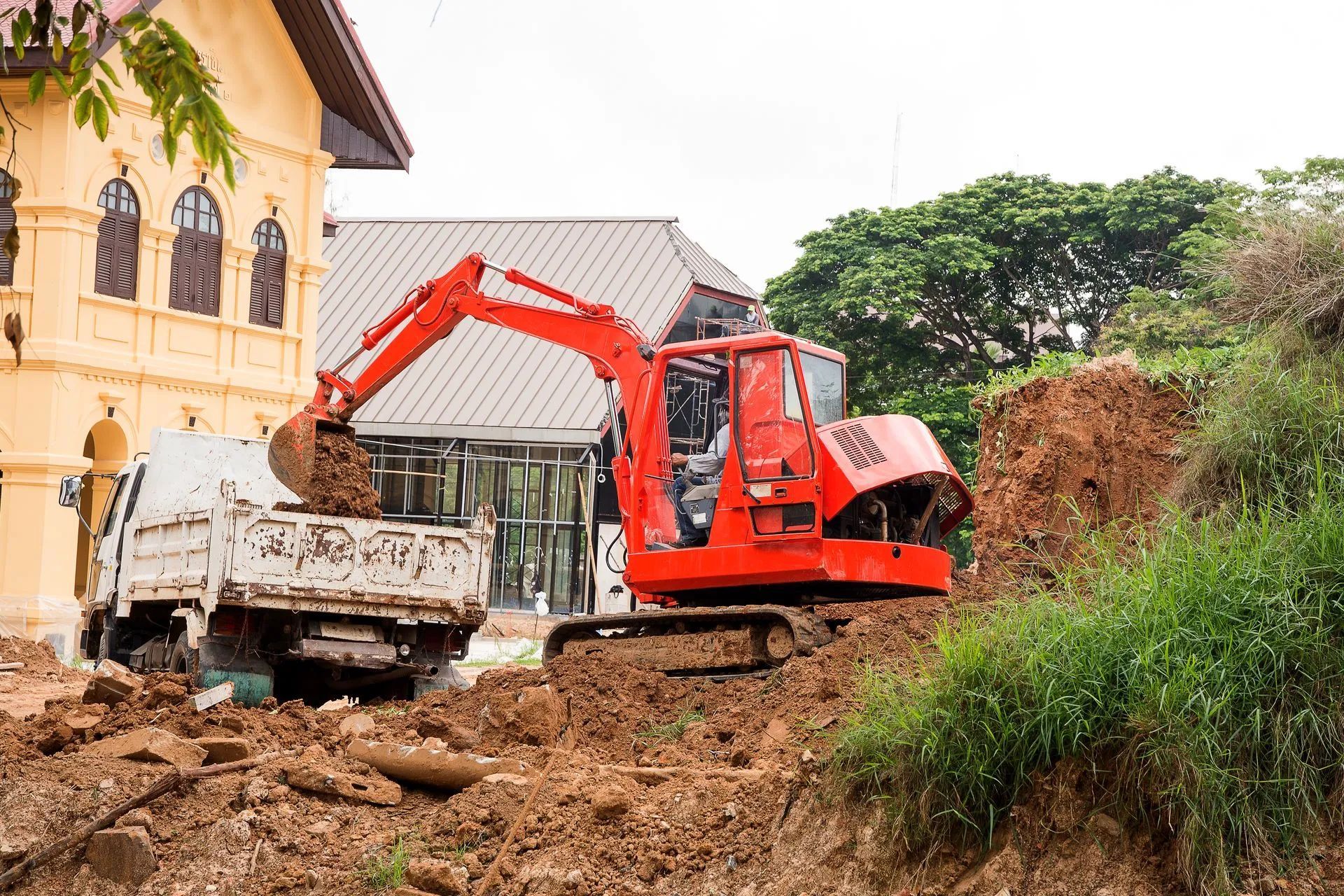 Red excavator loading debris into a dump truck beside a building on a muddy construction site