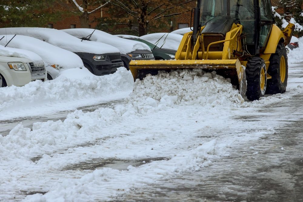 Yellow snowplow clearing deep snow beside parked cars in a snowy lot