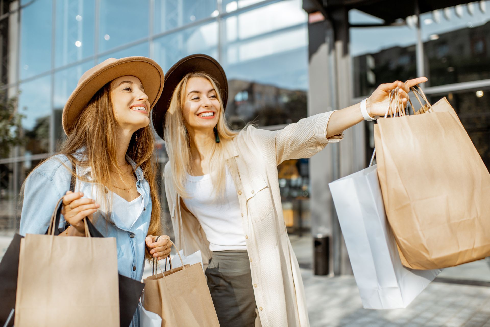 Two smiling young women wearing wide-brimmed hats stand outdoors on a sunny day holding several paper shopping bags. One of the women points into the distance, showing something to her friend in front of a modern glass-fronted building.