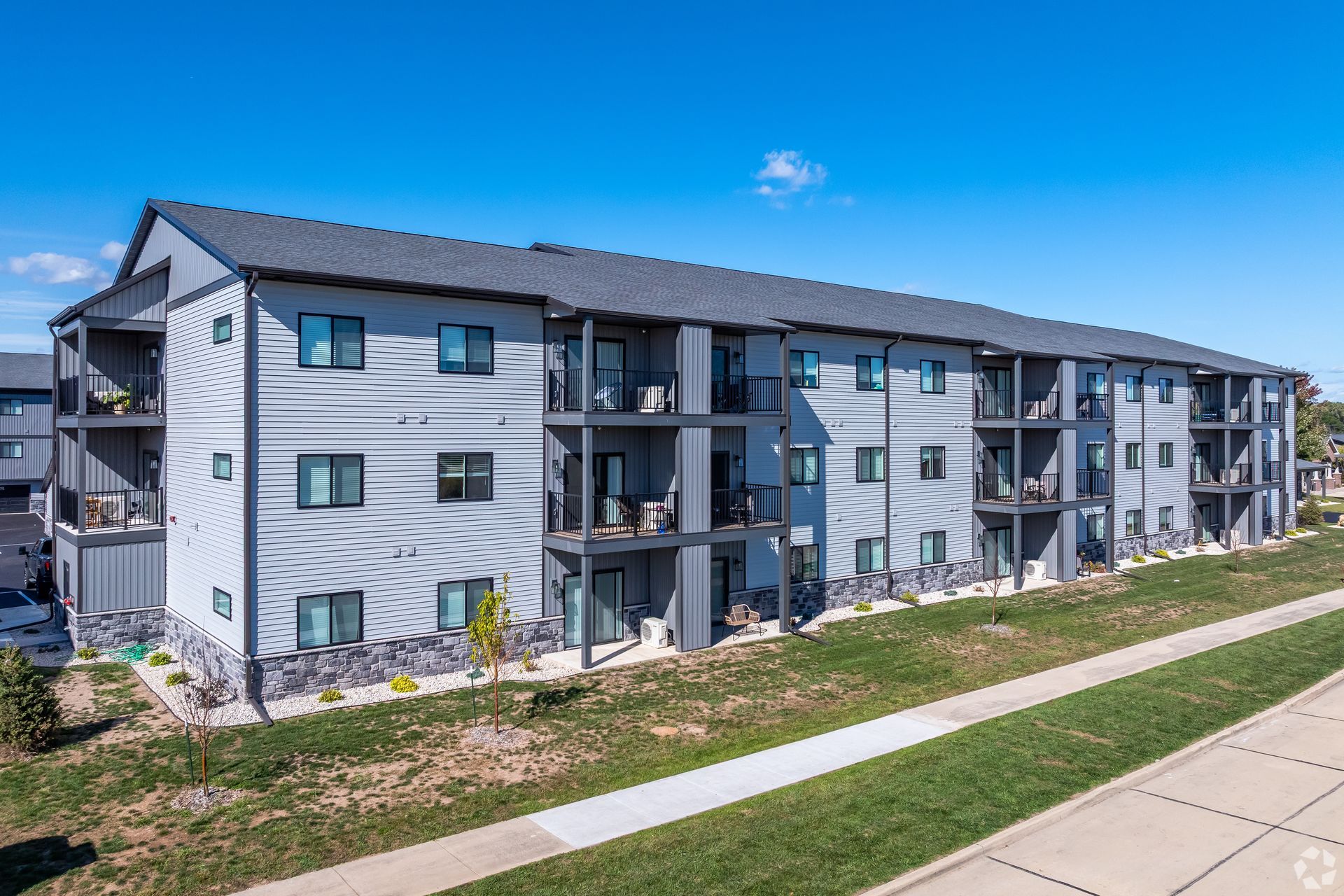 Three-story gray apartment building with balconies beside a sidewalk and landscaped lawn under a blue sky