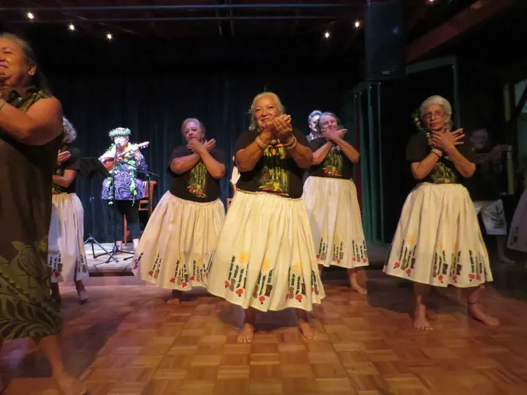 Dancers in white skirts and black tops performing on a wooden floor, audience in background.