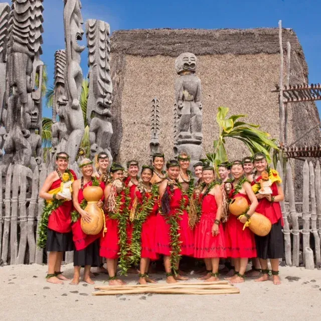 Group of people in red dresses and leis holding gourds, posing in front of carved wooden structures and a thatched-roof building.
