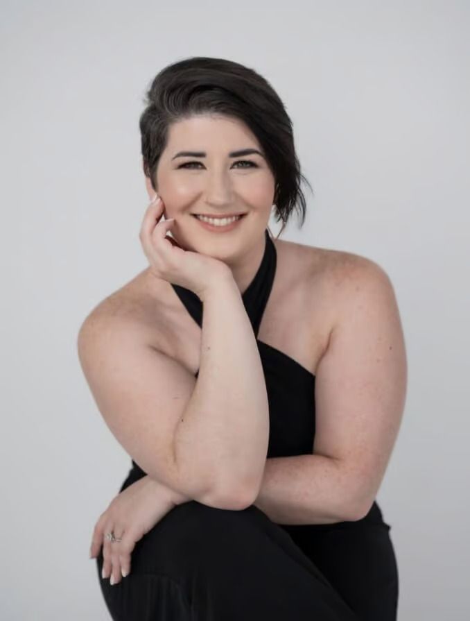 Woman with dark hair, smiling, wearing a black halter dress, resting her chin on her hand, against a white background.