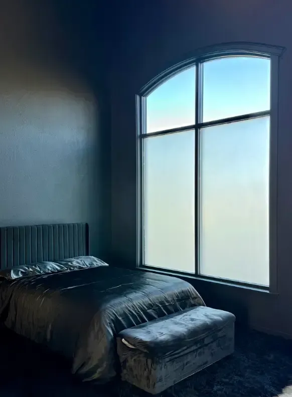 Bedroom with a tall arched window, bed, and bench. Soft gray tones and diffused light.