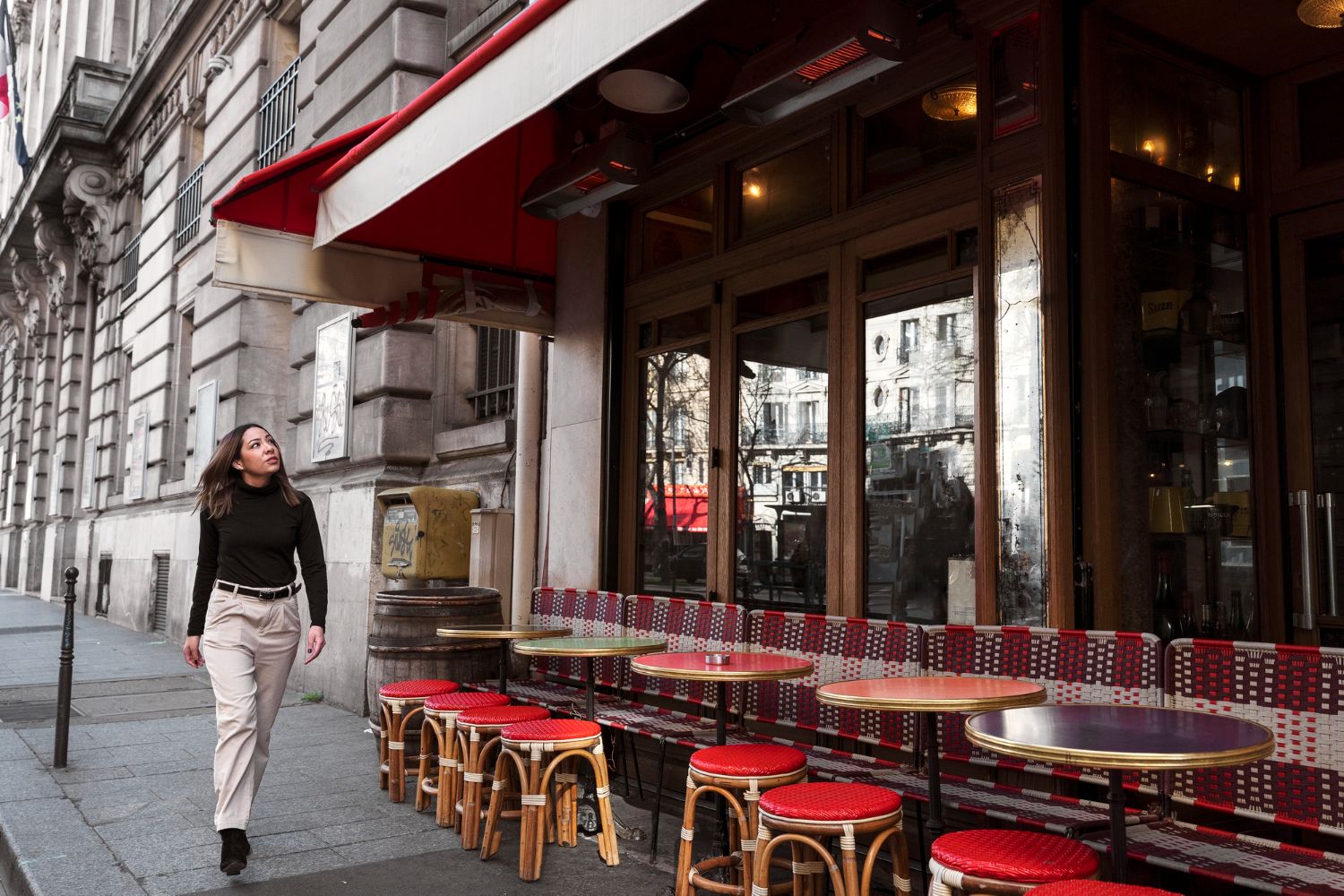 Woman walking past a coffee shop with red awning, tables, and chairs on a city street.