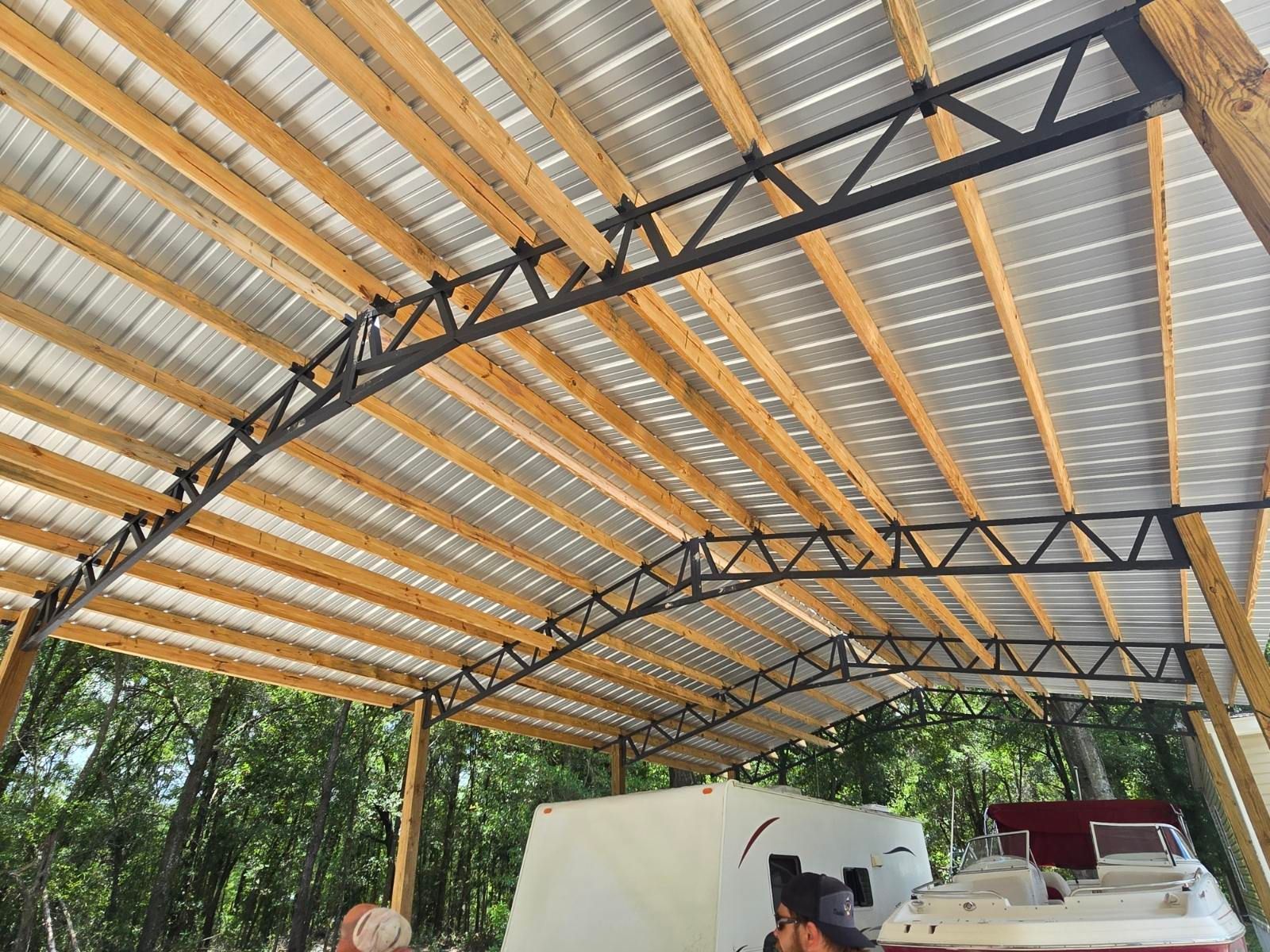 Covered open-air structure with metal roof trusses and parked machinery below, surrounded by trees