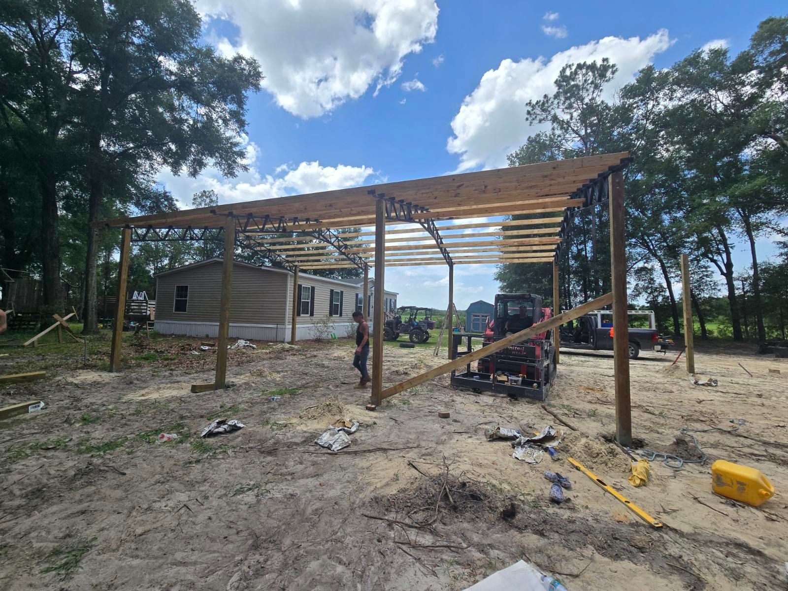 Partially built steel frame beside a small house, with construction materials on a dirt lot and trees nearby