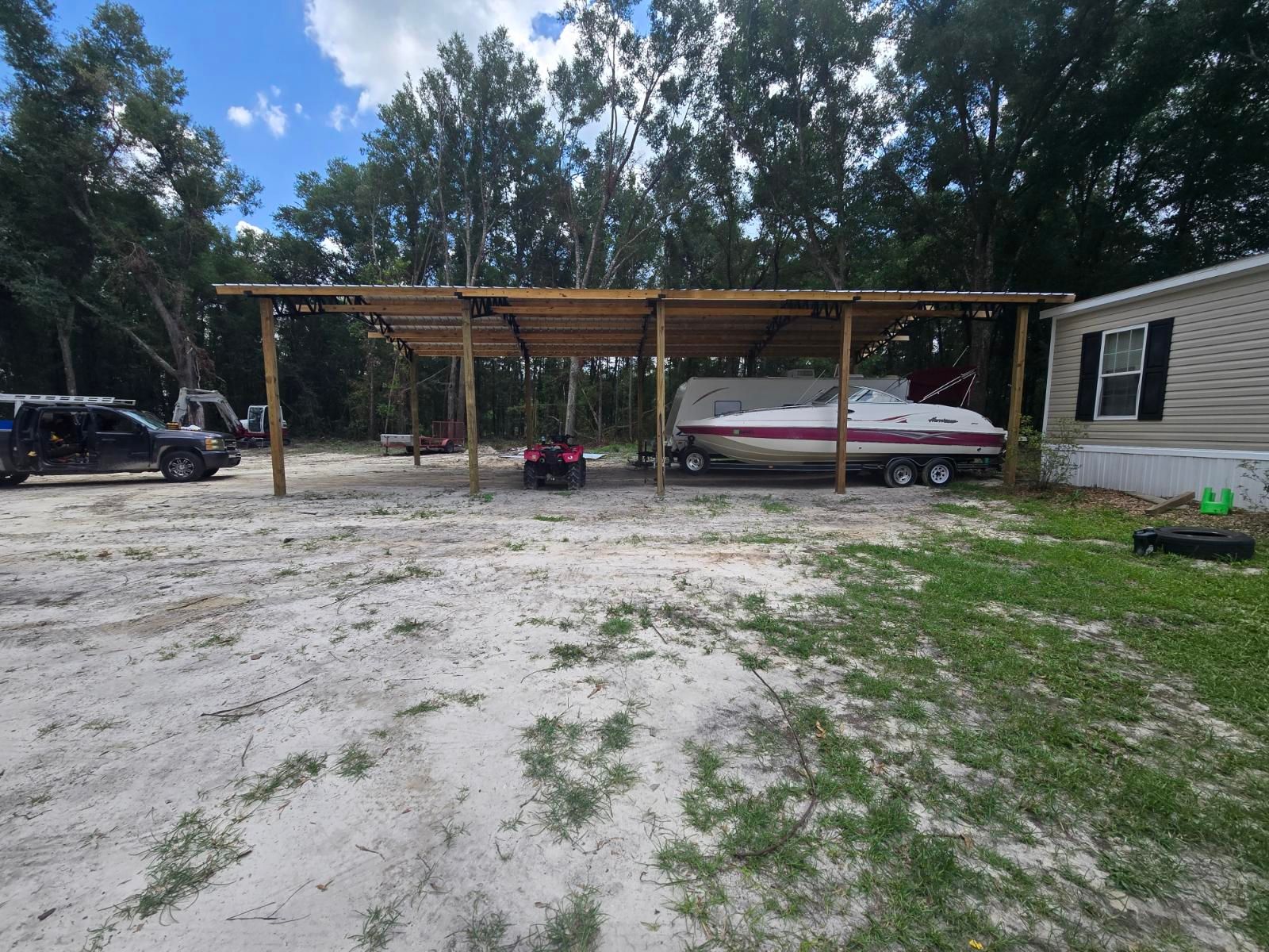 Rural yard with a wooden carport, red boat on a trailer, parked vehicles, and a small mobile home.