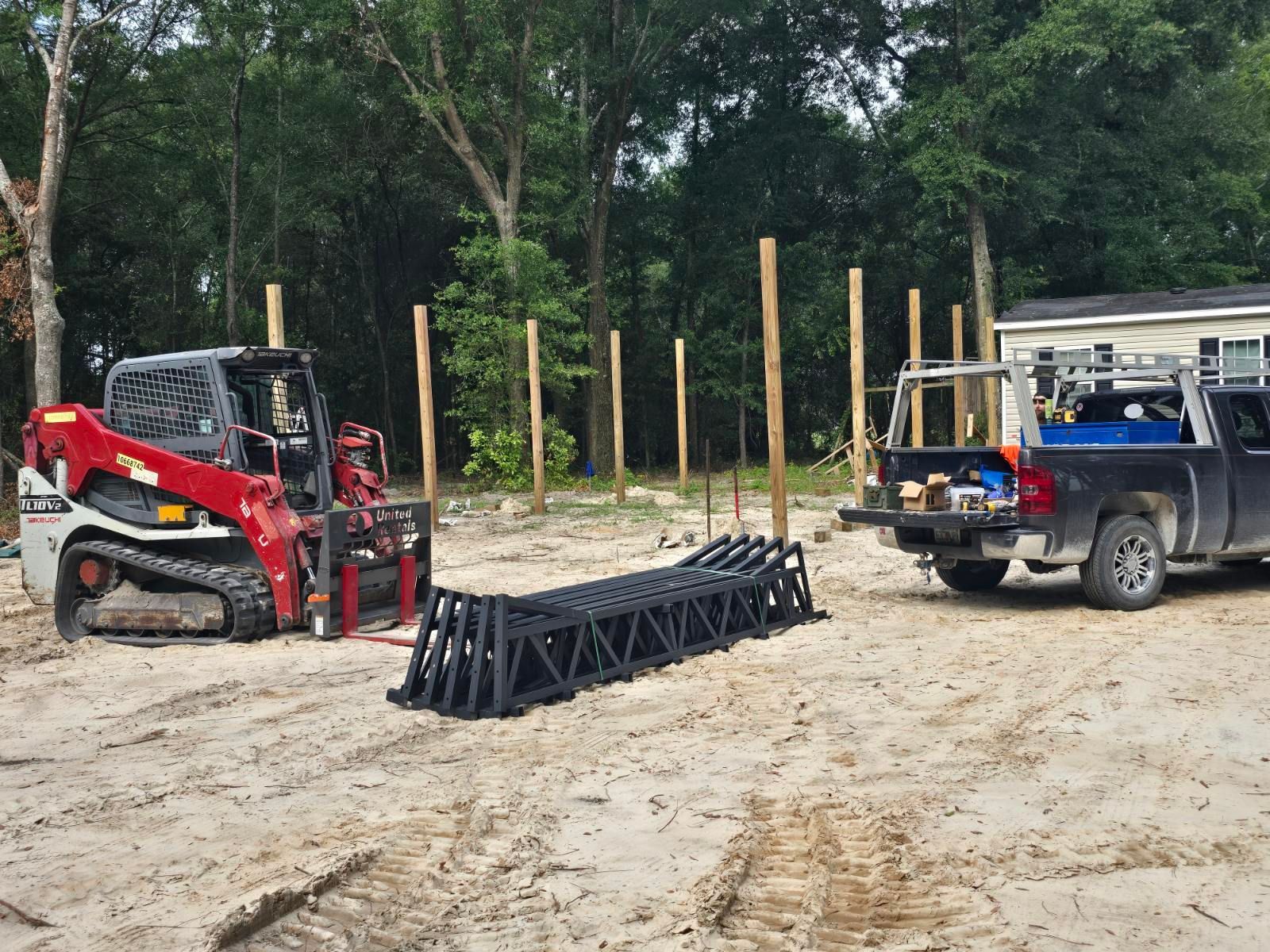 Red skid steer and blue pickup beside a metal rack on a dirt worksite with trees in the background