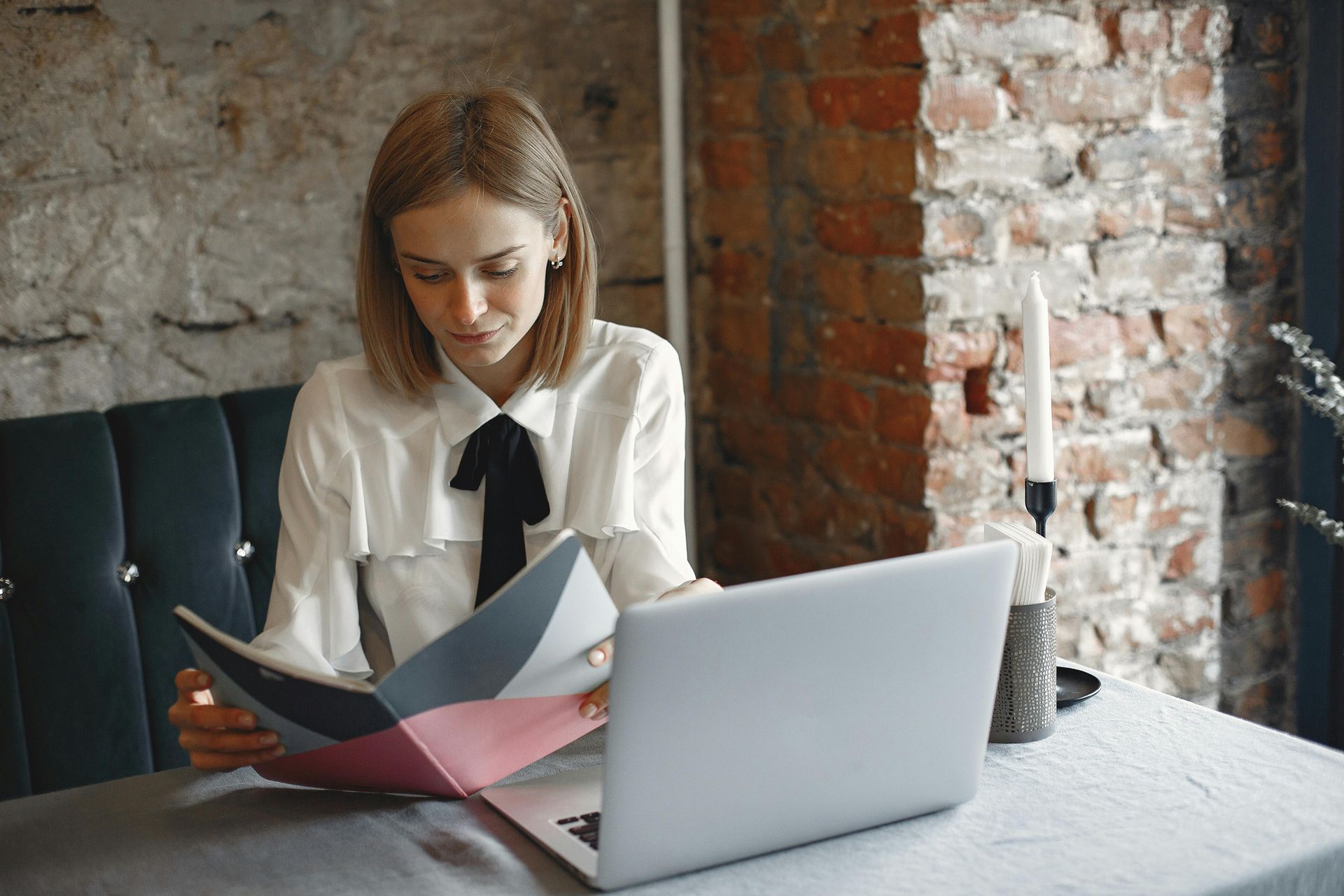 Woman reading a colorful folder at a table beside a laptop in a brick-walled café