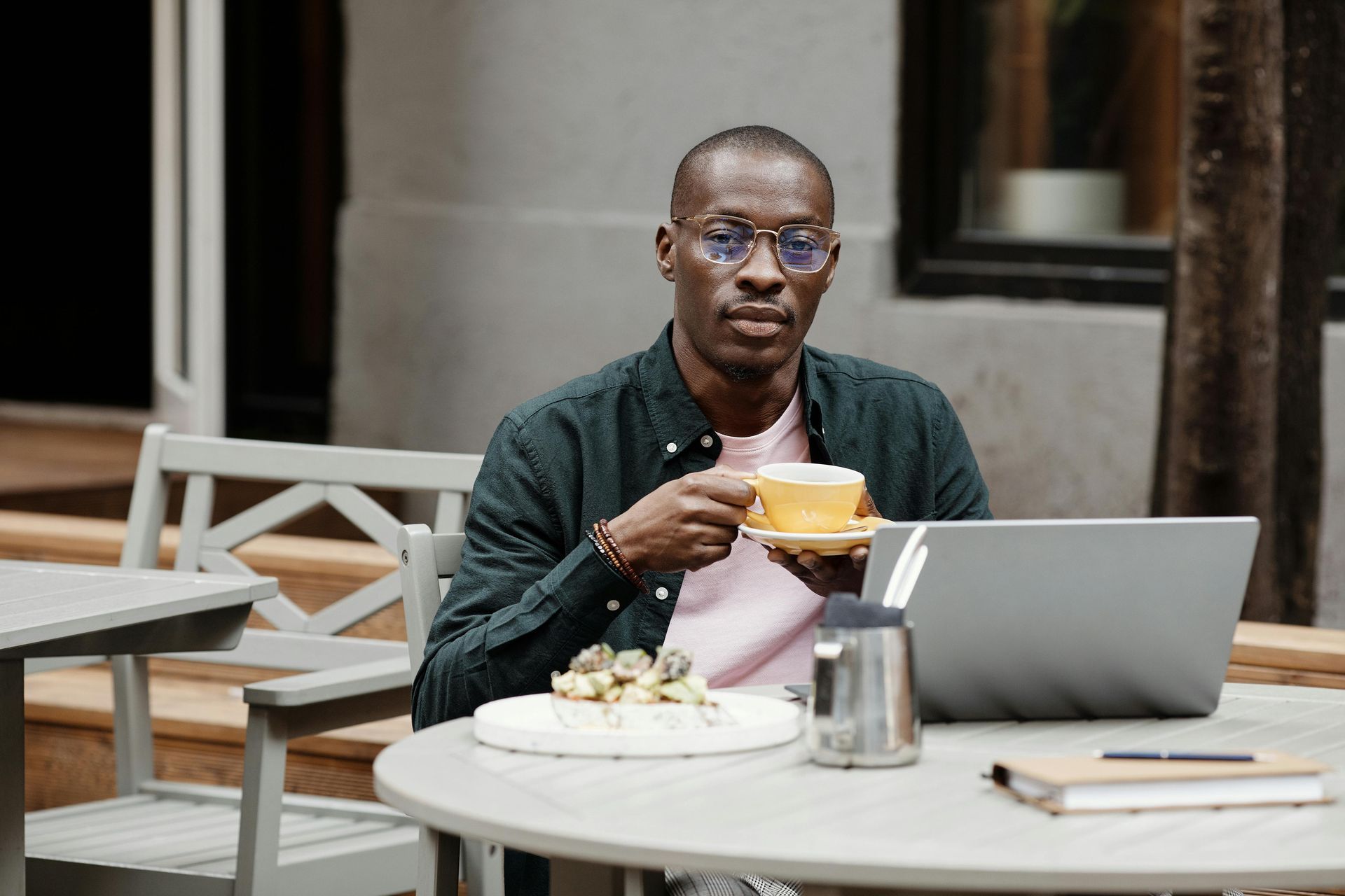 Person drinking from a yellow cup at a laptop in an outdoor café by a table with pastries.