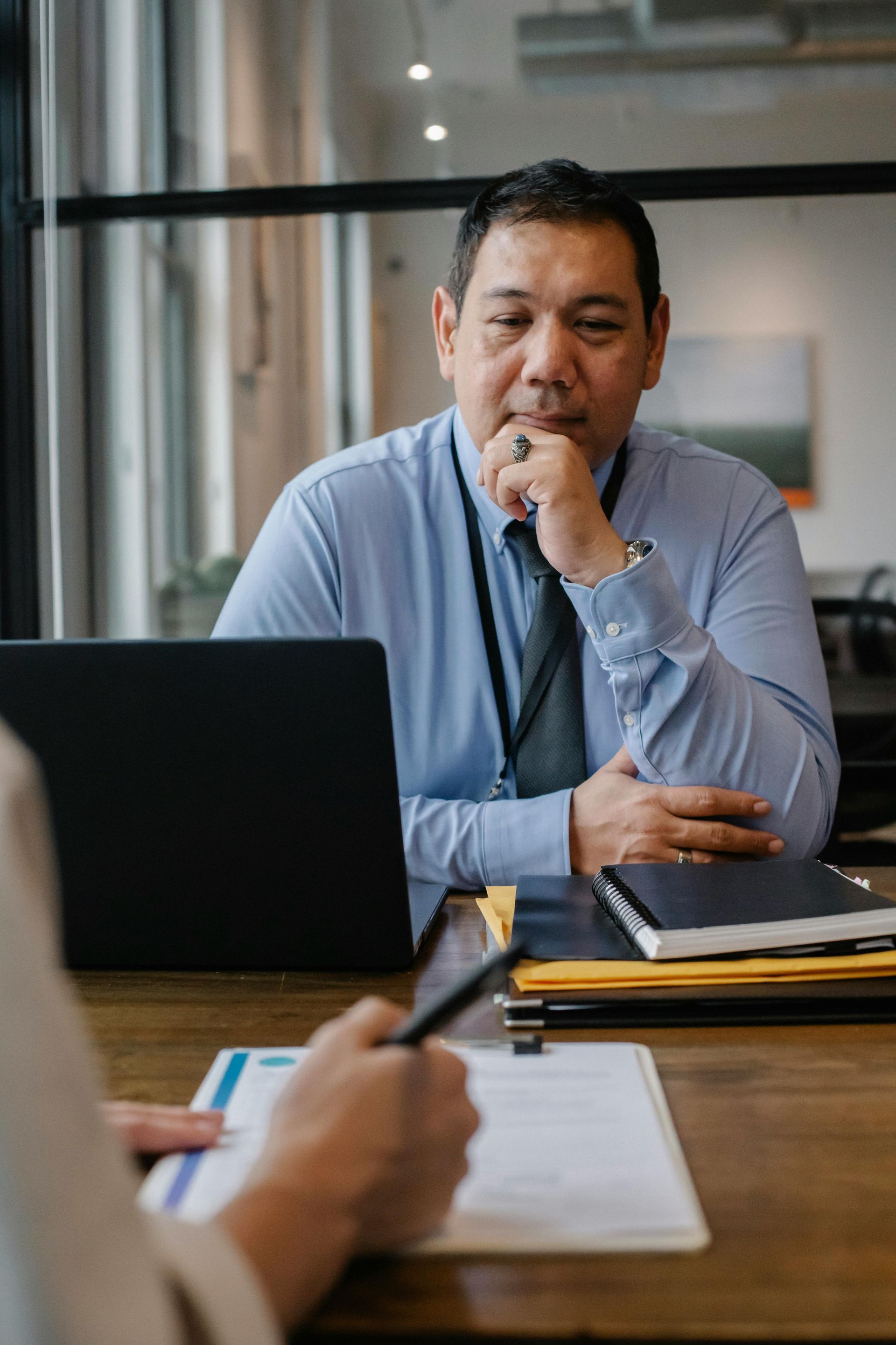 Man in a blue shirt thinking at a desk with a laptop in a meeting room
