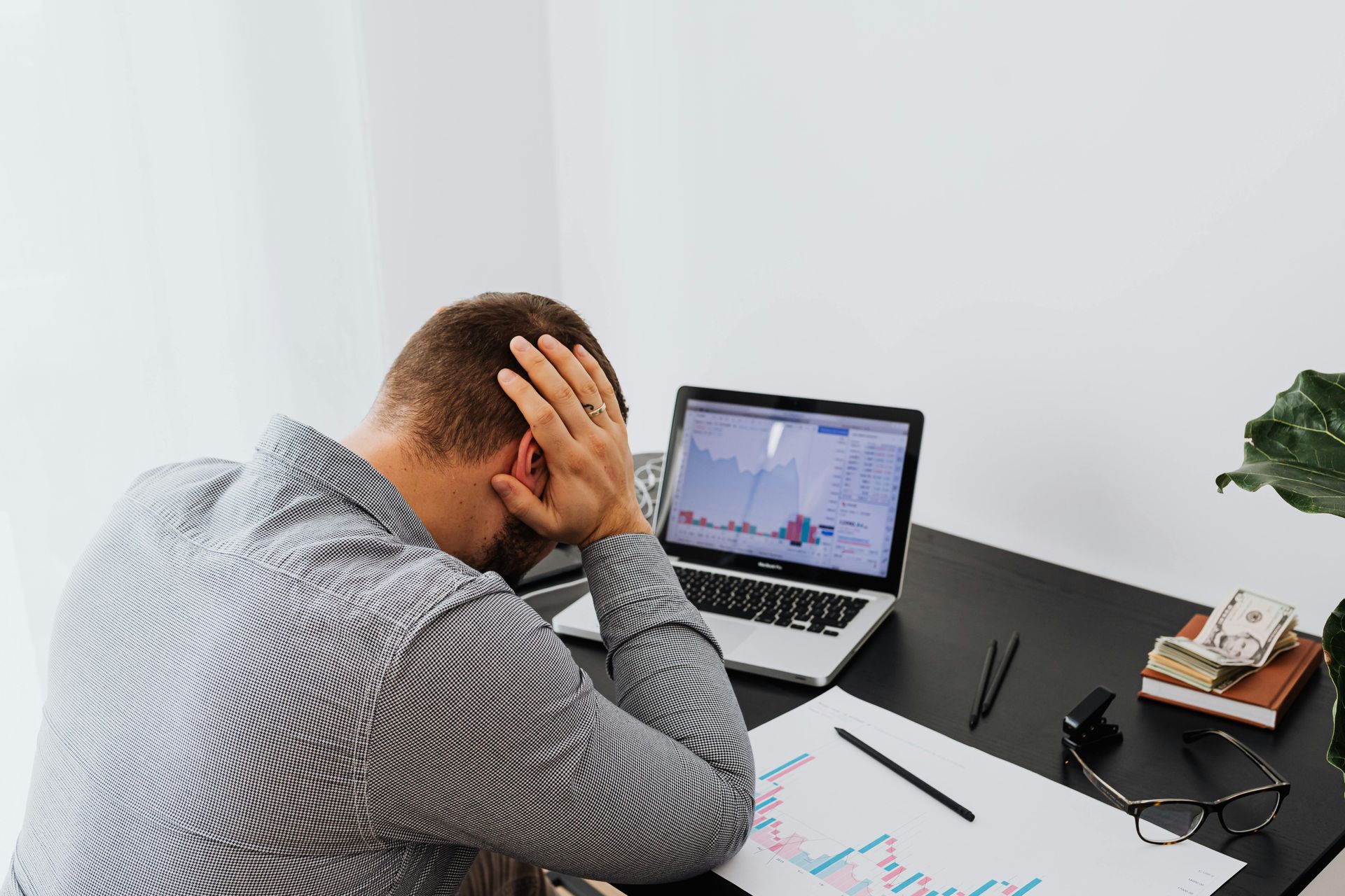 Person sitting at a desk, holding their head while looking at a laptop with charts and papers scattered nearby.
