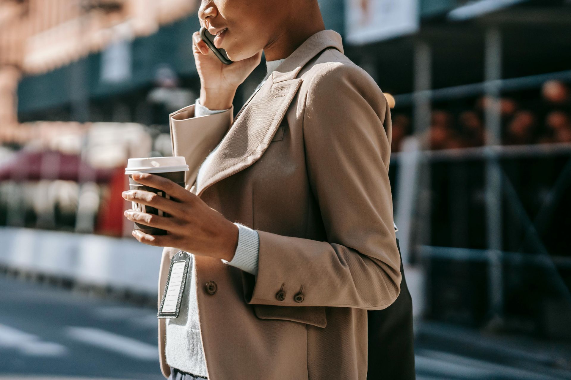 Person in a beige blazer on a phone call, holding a takeaway coffee on a city sidewalk.