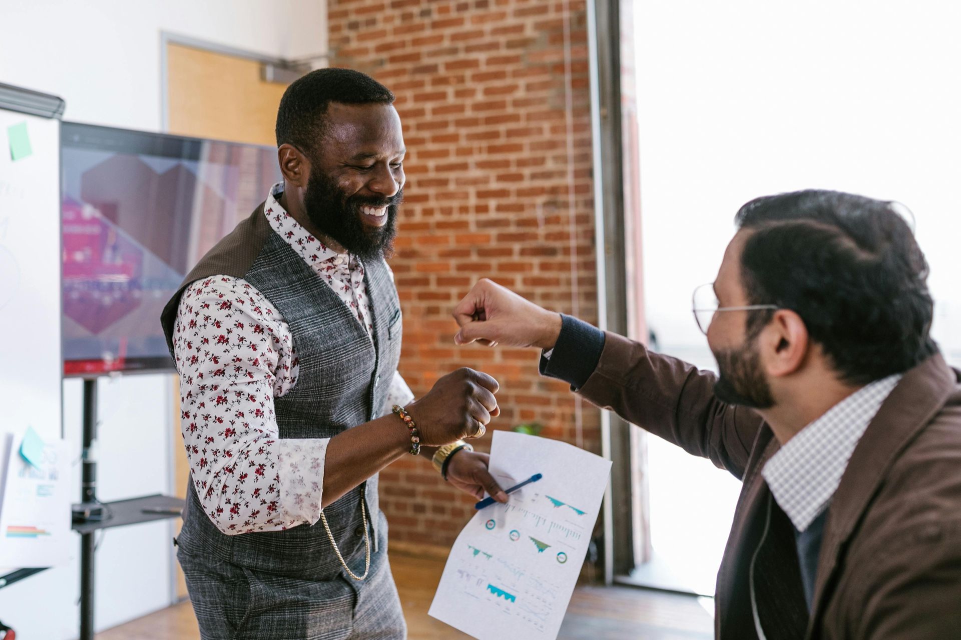 Two coworkers smiling and shaking hands in a bright office, one holding a chart.