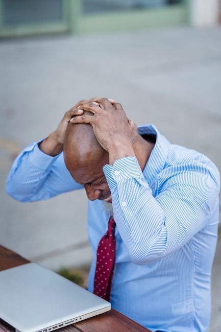 Man holding his head at a laptop, appearing stressed at a desk outdoors