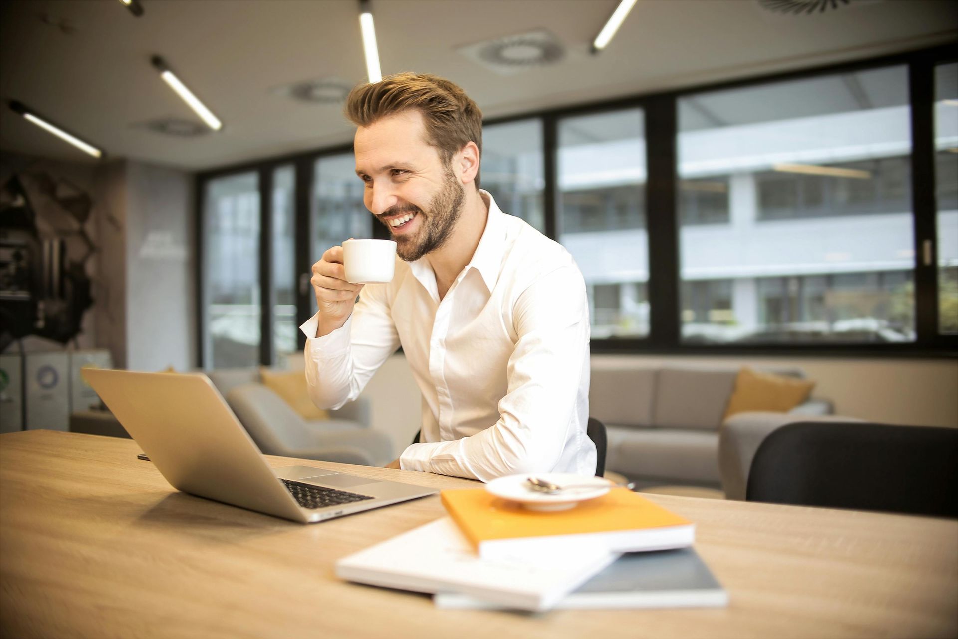 Man smiling with coffee beside a laptop at a desk in a bright office