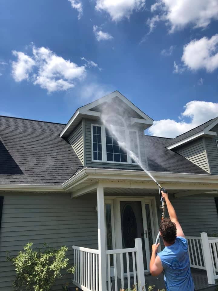 A person using a pressure washer to clean the exterior siding and window of a grey two-story house under a blue sky.