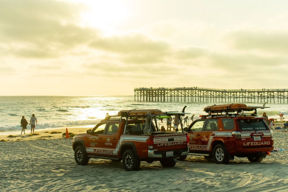 Lifeguard trucks parked on a beach near a pier under a cloudy sky.
