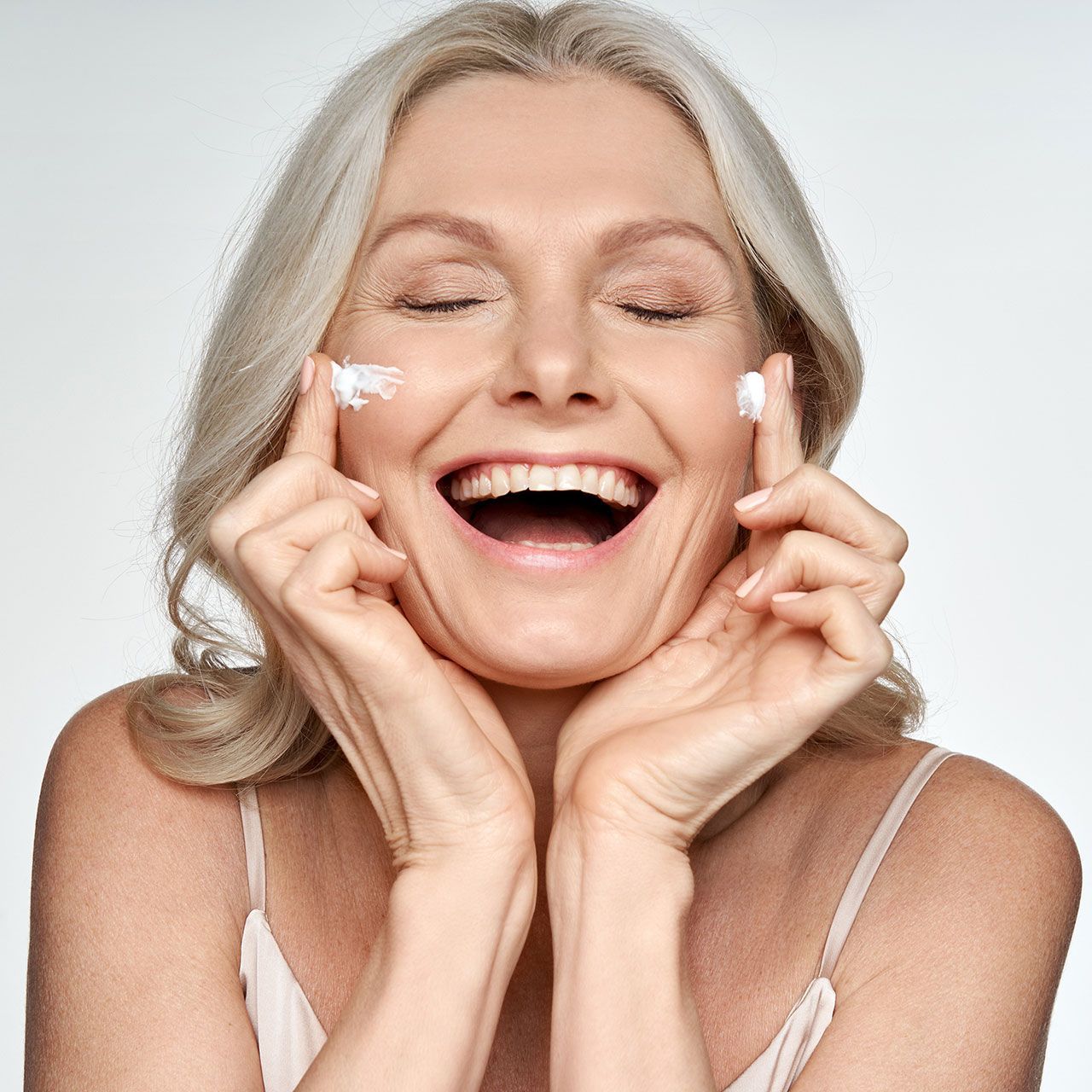 Happy funny 50s middle aged mature old blond woman applying facial cream on face laughing enjoying skin care beauty spa treatment creme isolated on white background. Mid age skincare concept