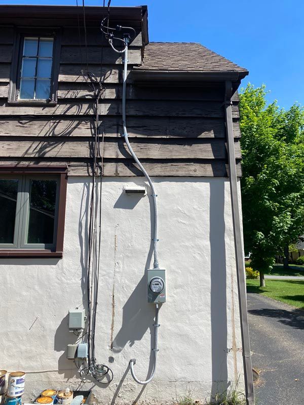 Side of a house with electrical meter, conduit, and utility wiring on a white and dark wood exterior