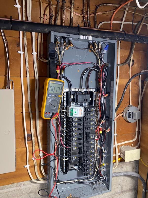 Open electrical breaker panel with multicolored wires in a basement wall, multimeter attached.