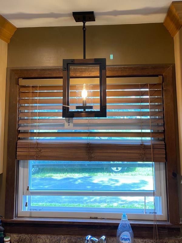 Kitchen window with wooden blinds and a black hanging light fixture above a sink ledge with a water bottle.