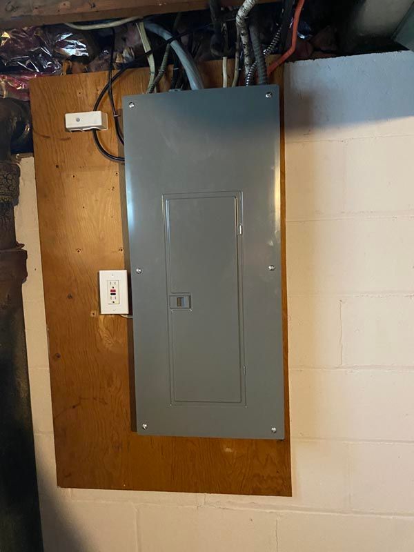 Gray electrical breaker panel mounted on a plywood wall with exposed wiring above it.