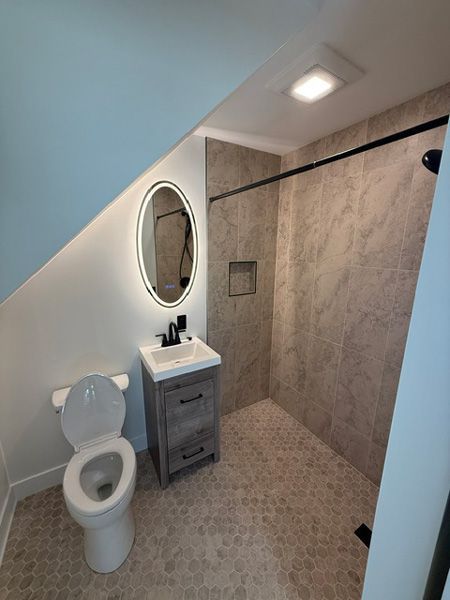 Small attic bathroom with toilet, vanity, oval mirror, and tiled shower under a sloped ceiling.