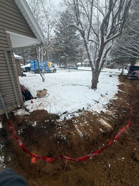 Snowy backyard with a fresh tree planting hole marked by red tape beside a house.