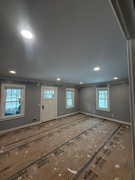 Empty gray room with recessed lights, white trim, windows, and bare subfloor exposed during renovation